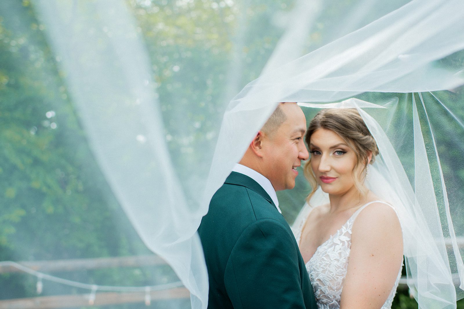 Melissa and Jonathan stand close together beneath her veil during outdoor portraits.